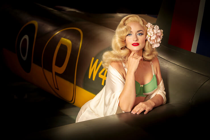 Did Derrier, a burlesque artist, poses for a photograph beside a warplane in an aircraft hanger.