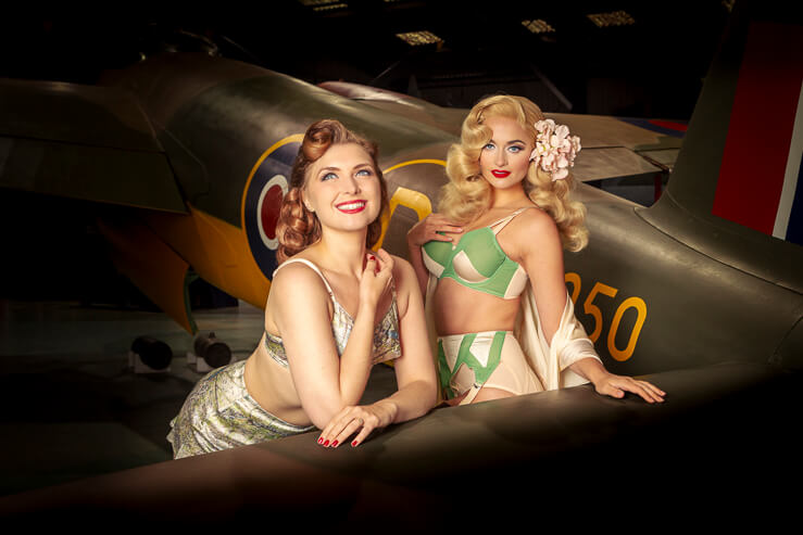 Felicity Furor and Didi Derrier,  burlesque models pose for a photograph by a Mosquito aircraft in an aircraft hanger.