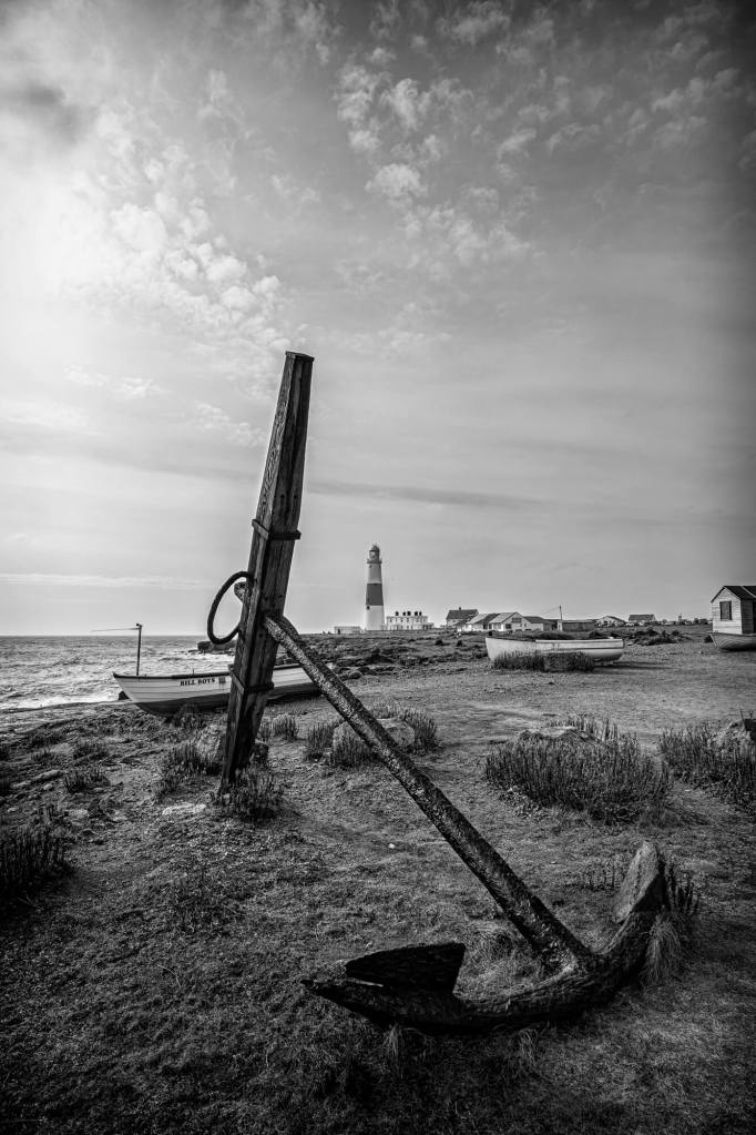 A large rusty old anchor on the shore, with Portland Bill Lighthouse behind.